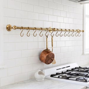 May include: A long, horizontal brass kitchen rail mounted to a wall, featuring multiple S-shaped hooks. A copper saucepan and a matching brass kitchen utensil hang from two of the hooks. The rail is secured with circular brass wall brackets.