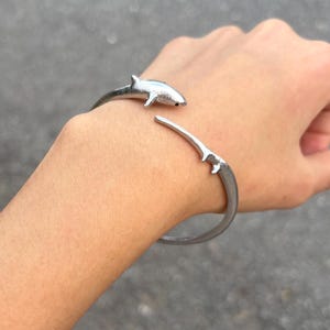 May include: A silver-toned metal cuff bracelet featuring a shark design. The bracelet is an open-ended style, with one end shaped like the head and body of a shark and the other end shaped like the shark's tail fin, creating a continuous wrap effect.