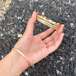 May include: Three gold-toned metal bangles featuring a continuous embossed floral pattern. Two of the bangles are stacked together, while the third bangle is worn separately on the wrist, all displaying a polished finish and a slim, circular design.