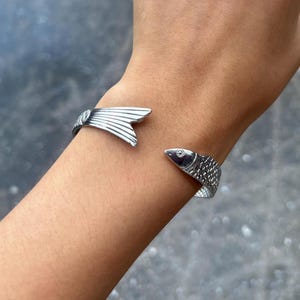 Puede incluir: Una pulsera de brazalete de metal plateado con un diseño de pez. Un extremo de la banda abierta tiene forma de cola de pez texturizada, mientras que el otro extremo es una cabeza de pez con escamas detalladas y un ojo visible en la superficie.