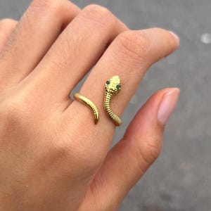 Puede incluir: Un anillo abierto de tono dorado con forma de serpiente enroscada, con un cuerpo texturizado y segmentado y dos pequeñas piedras verdes vibrantes como ojos. El anillo rodea el dedo, mostrando un diseño de cabeza detallado y una cola.