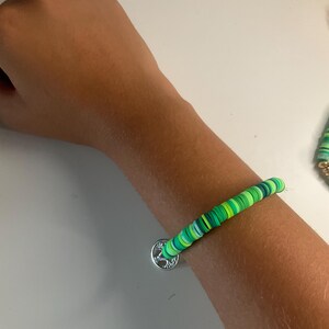 May include: A bracelet made of small, flat, circular clay beads in various shades of green, including lime, emerald, and forest green. The bracelet features a round, silver-toned metal charm with a detailed tree design hanging from the strand.