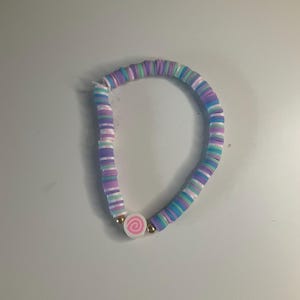 May include: A beaded bracelet featuring flat, circular clay discs in alternating shades of purple, light blue, white, and teal. The bracelet includes a single white circular charm with a pink spiral design, flanked by two small metallic gold beads.