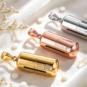Op de afbeelding: Drie cilindrische, pilvormige hangers in goud-, roségoud- en zilverkleurige tinten. Elke hanger heeft een metalen lus aan de bovenkant en is gegraveerd met een naam en een medisch symbool: Riley met een medische kit, Matilda met een caduceus en Isla.