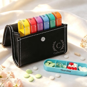 May include: A black leather-like pill organizer case with white stitching, featuring a personalized logo with the letter C and the name Catherine. It holds seven colorful daily pill containers. A separate light blue pill tray labeled Thursday sits nearby.