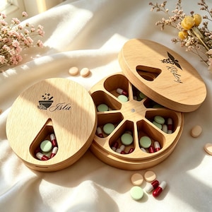 以下が含まれることがあります： 回転式の蓋が付いた2つの円形木製ピルケース。片方の蓋にはIslaという名前と乳鉢のアイコン、もう片方にはRileyという名前とカドゥケウスのシンボルが刻印されています。各容器には8つのくさび形のコンパートメントが備わっています。
