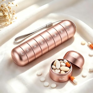 Op de afbeelding: Een roségouden cilindrische pillendoos met zeven stapelbare compartimenten gelabeld met de dagen van de week van SUN tot SAT. De houder is voorzien van een grijs polsbandje en de merknaam Ruby op de bovenkant. Eén compartiment staat open.