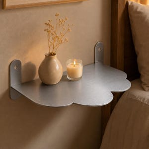 May include: A wall-mounted metal shelf with a brushed silver finish and a scalloped front edge. The shelf features two vertical mounting brackets with circular screw holes. A beige ceramic vase with dried flowers and a lit candle sit on the surface.