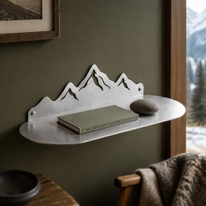 Könnte beinhalten: Ein silbernes Wandregal aus Metall mit einer dekorativen Rückwand in Form einer Bergkette. Das Regal hat eine glatte, abgerundete Ablagefläche, auf der ein grünes Buch mit dem Titel The Alpine Path und ein kleiner, glatter grauer Stein liegen.