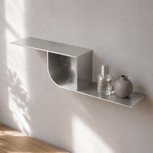 May include: A brushed metal wall shelf featuring a flat top surface, a curved open compartment, and a lower flat surface. A clear glass bottle with a silver cap and a small, round, matte gray ceramic vase sit on the lower shelf section.
