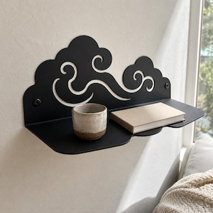 May include: A matte black metal wall shelf featuring a decorative back panel with a cutout cloud pattern. The shelf holds a small, speckled ceramic cup and a rectangular book with a plain tan cover. The unit is mounted with two visible screws.
