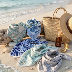 May include: A collection of five square cotton scarves featuring various blue, green, and grey floral and paisley patterns. The scarves are arranged alongside a woven straw tote bag, a wide-brimmed straw hat, a bottle of SPF oil, and a gold compass.