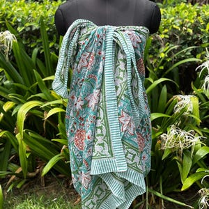 May include: A sarong featuring a teal and white floral pattern with red accents. The fabric displays a repeating botanical print with large blossoms and leaves, finished with a striped border along the edges, draped over a black mannequin torso.