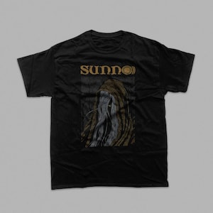 May include: A black short-sleeve t-shirt featuring the gold text SUNNO))) above a graphic illustration of a hooded figure with long flowing hair. The figure is depicted in shades of grey and gold, set against a dark, textured background.
