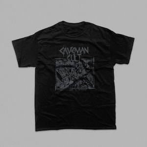 May include: A black short-sleeve t-shirt featuring a grey graphic print on the chest. The design includes the text "CAVEMAN CULT" in a stylized font above a rectangular illustration of a battlefield scene with the text "SAVAGE WAR IS DESTINY".