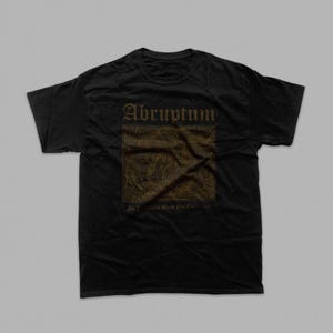 以下が含まれることがあります： 黒の半袖Tシャツ。ゴシック体のフォントで「Abruptum」と書かれ、その下に長方形の茶色のグラフィックイラストが配置されている。デザインには複雑な線画が含まれ、下部には小さなゴシック体のフォントで追加のテキストが記されている。
