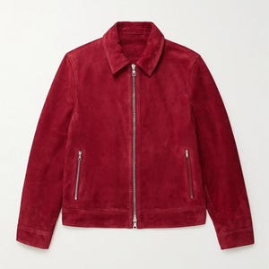 Puede incluir: Una chaqueta de ante de color rojo intenso con cuello clásico, mangas largas con puños abotonados y un cierre de cremallera metálica plateada en el centro. La prenda incluye dos bolsillos verticales con cremallera en la parte inferior.