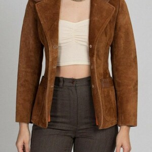 Può includere: Un blazer in camoscio marrone con revers dentellato, maniche lunghe e chiusura frontale con bottoni. La giacca presenta due tasche anteriori con finiture in pelle lungo il colletto e i bordi delle tasche, indossata sopra un top corto color crema.