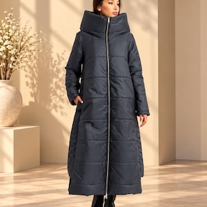 Peut inclure: Un long manteau doudoune gris anthracite foncé doté d'un col haut surdimensionné, d'une fermeture éclair intégrale sur le devant et de surpiqûres horizontales. Le manteau descend jusqu'au bas du mollet et est porté avec des bottines noires.