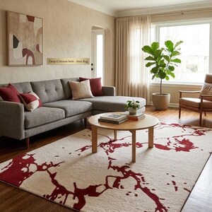 Puede incluir: Una alfombra rectangular con una base de color crema y un patr&oacute;n abstracto de color rojo oscuro en forma de vetas extendido por la superficie. La alfombra est&aacute; colocada sobre un suelo de madera en una habitaci&oacute;n con un sof&aacute; gris y una mesa.