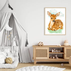 May include: A framed wall art print featuring a watercolor illustration of a brown fawn with white spots resting on a patch of green grass against a light blue sky background. The artwork is displayed in a simple light-colored wooden frame.