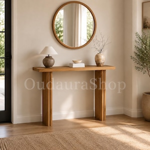 May include: A light brown wooden console table with vertical fluted legs. On the table sits a lamp with a white shade and a textured base, a small white bowl on a stack of books, and a vase with thin branches. A round mirror hangs above.