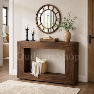 May include: A rectangular wooden console table with a dark brown finish and a hollow center. On top are two black candle holders with white candles, a woven tray with books, and a ceramic vase with greenery. A woven basket with a cloth sits inside.