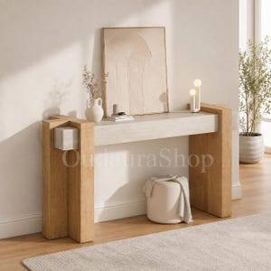 May include: A light wood console table featuring thick, block-style legs and a rectangular top. The table has a light-colored horizontal beam connecting the legs. A white vase, a framed textured art piece, and a small lamp sit on the surface.