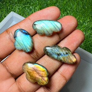 May include: Four small, polished labradorite stones carved into cloud shapes. Each stone displays iridescent flashes of blue, gold, and green light across its smooth, reflective surface, showcasing the natural mineral properties of the gemstone.