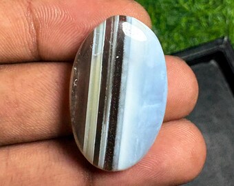Può includere: Una gemma a cabochon di forma ovale con distinte bande verticali di colore. La pietra presenta una superficie liscia e lucida con strisce alternate di marrone scuro, marrone chiaro traslucido e una sezione solida bianco latte opaco.