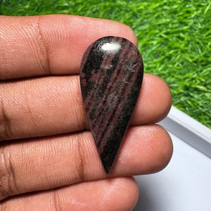 May include: A teardrop-shaped cabochon gemstone featuring a polished surface with a mix of deep burgundy, dark brown, and black streaks. The stone has a smooth, rounded top and a pointed bottom, displaying a natural, varied color pattern throughout.