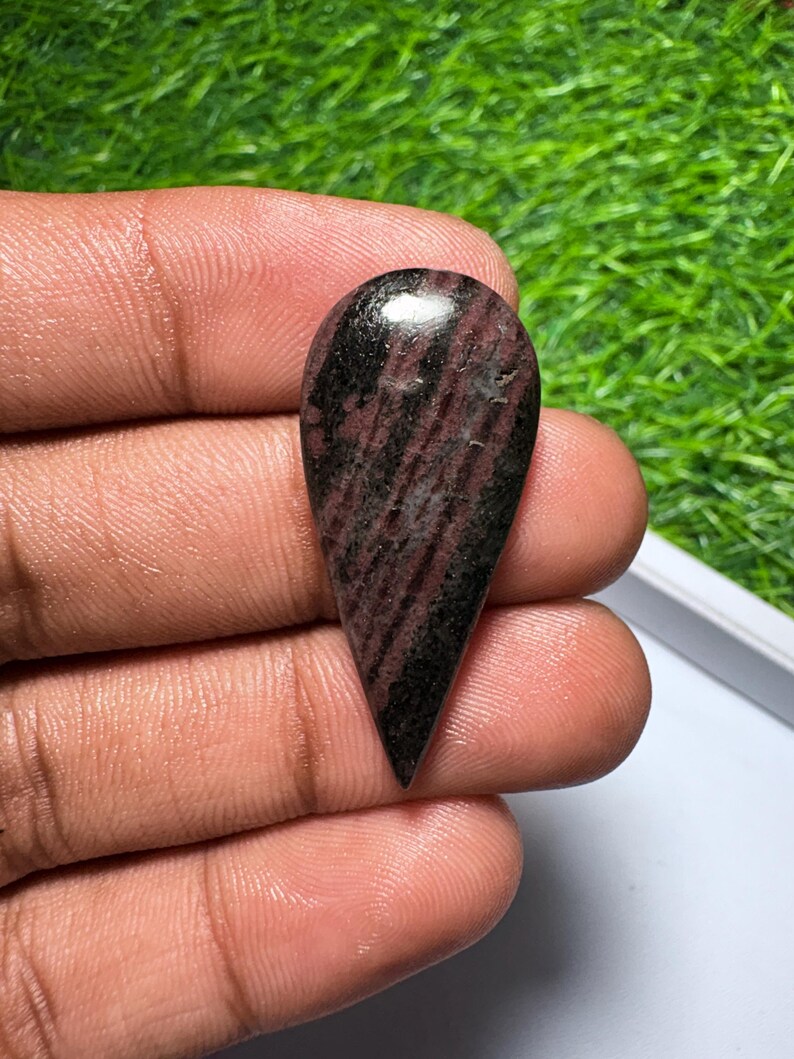 May include: A teardrop-shaped cabochon gemstone featuring a polished surface with a mix of deep burgundy, dark brown, and black streaks. The stone has a smooth, rounded top and a pointed bottom, displaying a natural, varied color pattern throughout.