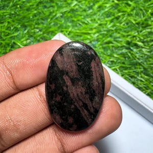 Peut inclure: Une pierre pr&eacute;cieuse polie de forme ovale pr&eacute;sentant un motif sombre et tachet&eacute; de teintes noir profond et rouge brun&acirc;tre. La pierre poss&egrave;de une surface lisse et r&eacute;fl&eacute;chissante avec une finition brillante, r&eacute;v&eacute;lant des inclusions min&eacute;rales.