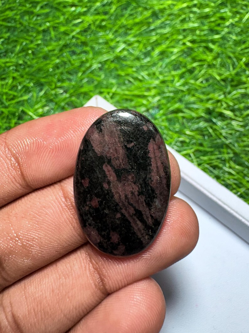Peut inclure: Une pierre pr&eacute;cieuse polie de forme ovale pr&eacute;sentant un motif sombre et tachet&eacute; de teintes noir profond et rouge brun&acirc;tre. La pierre poss&egrave;de une surface lisse et r&eacute;fl&eacute;chissante avec une finition brillante, r&eacute;v&eacute;lant des inclusions min&eacute;rales.