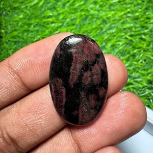 Pode incluir: Uma pedra preciosa caboch&atilde;o de formato oval com uma superf&iacute;cie polida e um padr&atilde;o manchado de cores bord&ocirc; profundo e preto. A pedra tem um topo liso e arredondado com um acabamento brilhante que reflete a luz na superf&iacute;cie.