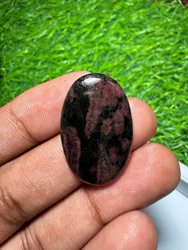 Pode incluir: Uma pedra preciosa caboch&atilde;o de formato oval com uma superf&iacute;cie polida e um padr&atilde;o manchado de cores bord&ocirc; profundo e preto. A pedra tem um topo liso e arredondado com um acabamento brilhante que reflete a luz na superf&iacute;cie.