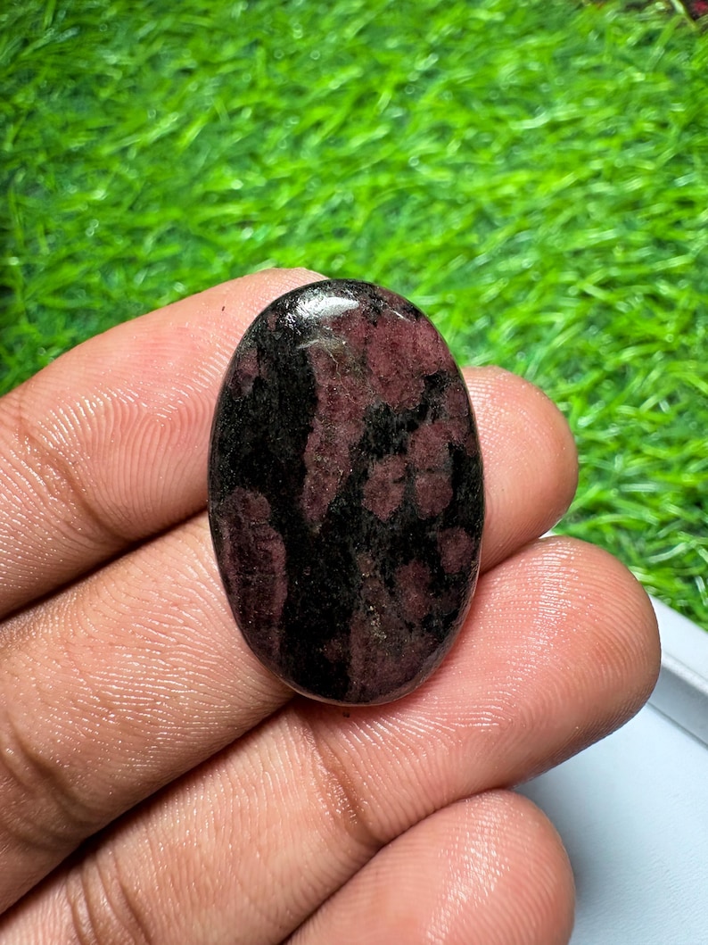 Pode incluir: Uma pedra preciosa caboch&atilde;o de formato oval com uma superf&iacute;cie polida apresentando um padr&atilde;o manchado de vermelho bord&ocirc; profundo e inclus&otilde;es minerais pretas escuras, com uma c&uacute;pula lisa e acabamento brilhante.