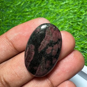 Pode incluir: Uma pedra preciosa polida de formato oval com uma superf&iacute;cie lisa e um padr&atilde;o manchado de vermelho bord&ocirc; profundo e preto s&oacute;lido. A pedra tem um acabamento brilhante que reflete a luz, destacando o contraste entre os tons escuros e vermelhos.