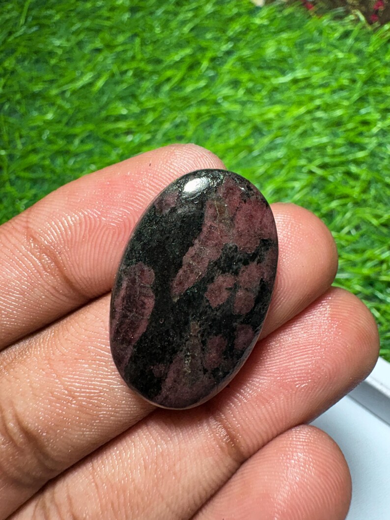 Pode incluir: Uma pedra preciosa polida de formato oval com uma superf&iacute;cie lisa e um padr&atilde;o manchado de vermelho bord&ocirc; profundo e preto s&oacute;lido. A pedra tem um acabamento brilhante que reflete a luz, destacando o contraste entre os tons escuros e vermelhos.