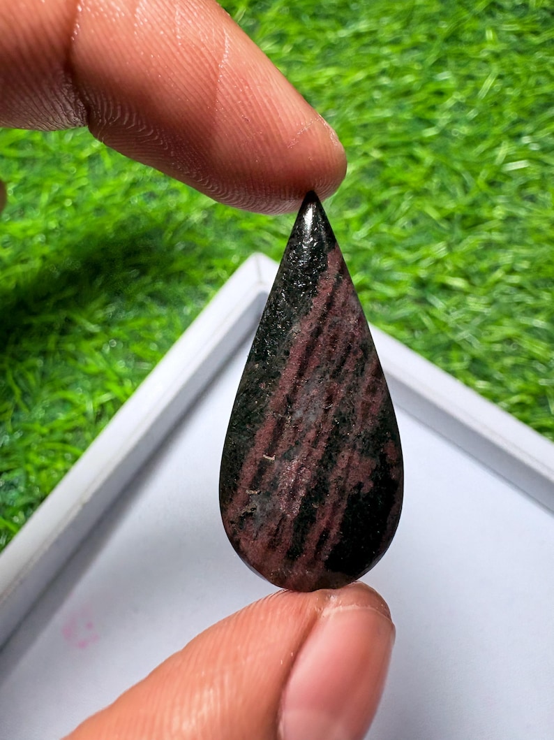 May include: A teardrop-shaped gemstone featuring a polished surface with a distinct pattern of deep black and dark maroon streaks running vertically throughout the stone, showcasing a smooth, reflective finish and a pointed top.