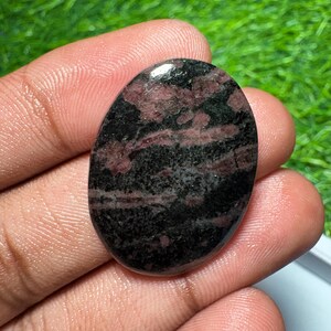 Pu&ograve; includere: Una pietra preziosa lucidata di forma ovale con una superficie liscia e un motivo maculato nei colori nero profondo e rosso bordeaux scuro. La pietra ha una finitura lucida che riflette la luce, mettendo in risalto le inclusioni minerali irregolari.