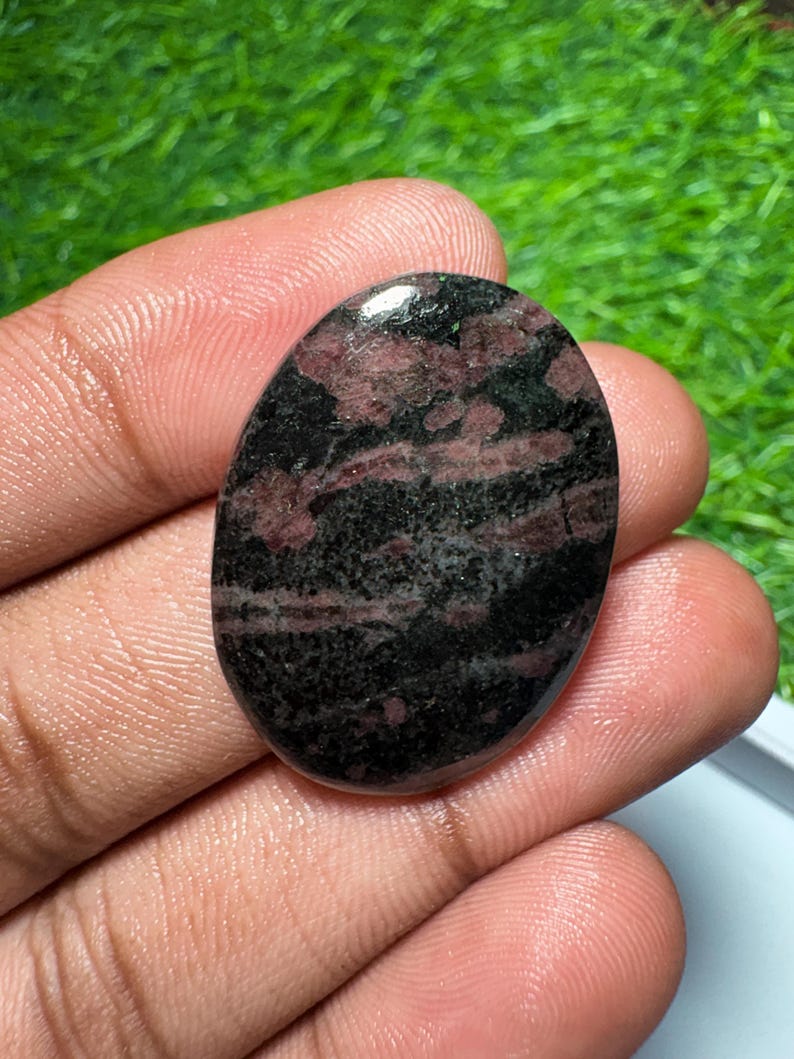 Pu&ograve; includere: Una pietra preziosa lucidata di forma ovale con una superficie liscia e un motivo maculato nei colori nero profondo e rosso bordeaux scuro. La pietra ha una finitura lucida che riflette la luce, mettendo in risalto le inclusioni minerali irregolari.