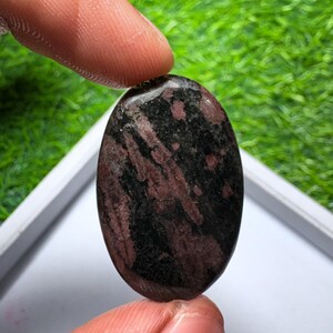 Peut inclure: Une pierre pr&eacute;cieuse cabochon de forme ovale pr&eacute;sentant une surface polie avec un motif tachet&eacute; de noir profond et de rouge bordeaux fonc&eacute;. La pierre a une texture lisse et arrondie ainsi qu'une finition brillante.