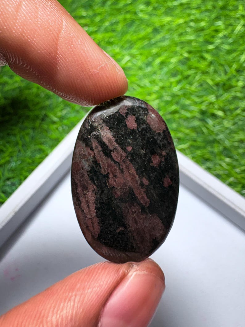 Peut inclure: Une pierre pr&eacute;cieuse cabochon de forme ovale pr&eacute;sentant une surface polie avec un motif tachet&eacute; de noir profond et de rouge bordeaux fonc&eacute;. La pierre a une texture lisse et arrondie ainsi qu'une finition brillante.