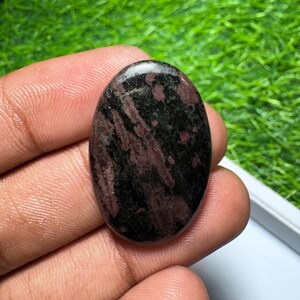 Peut inclure: Une pierre pr&eacute;cieuse polie de forme ovale pr&eacute;sentant une base noire fonc&eacute;e avec des stries et des motifs tachet&eacute;s bordeaux et marron profond sur toute la surface lisse et r&eacute;fl&eacute;chissante de la pierre.