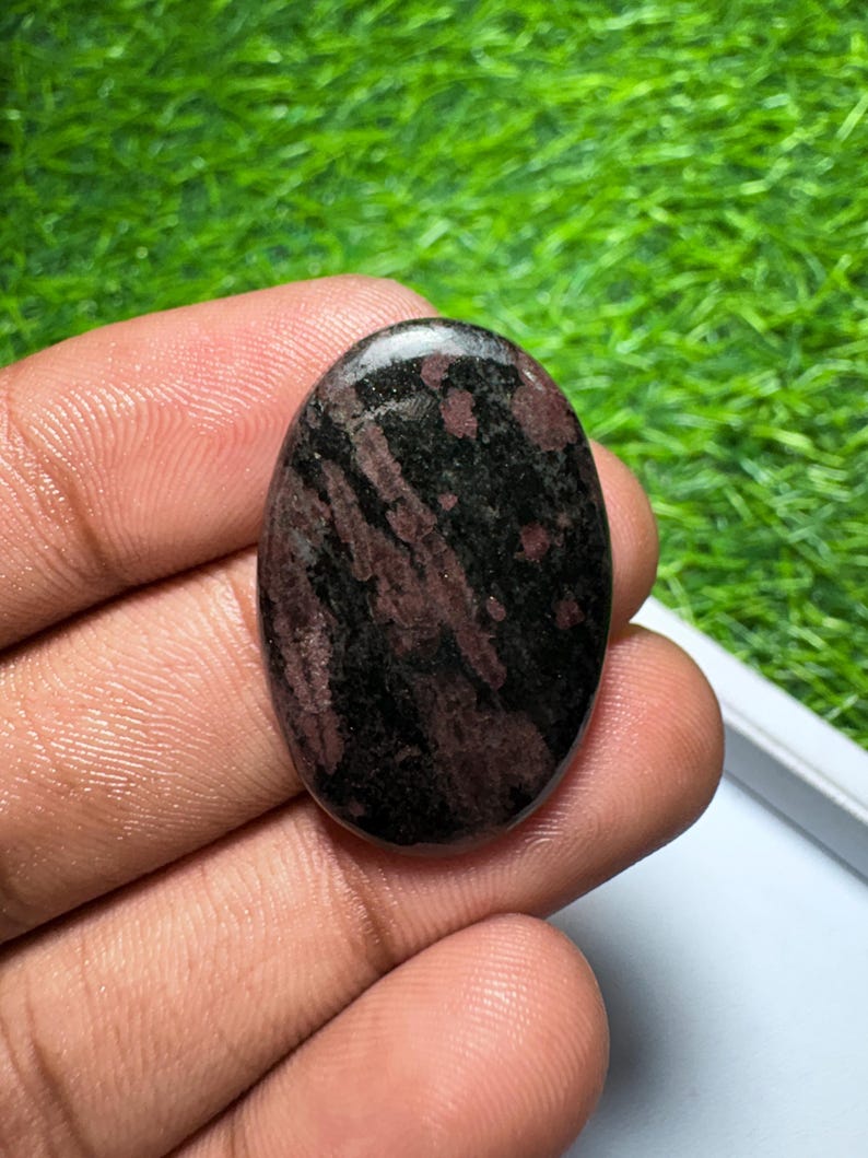 Peut inclure: Une pierre pr&eacute;cieuse polie de forme ovale pr&eacute;sentant une base noire fonc&eacute;e avec des stries et des motifs tachet&eacute;s bordeaux et marron profond sur toute la surface lisse et r&eacute;fl&eacute;chissante de la pierre.