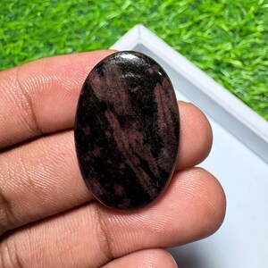 Peut inclure: Une pierre pr&eacute;cieuse polie de forme ovale pr&eacute;sentant un motif tachet&eacute; rouge fonc&eacute; et noir. La pierre poss&egrave;de une surface lisse et r&eacute;fl&eacute;chissante avec des stries et des taches sombres irr&eacute;guli&egrave;res r&eacute;parties sur sa base rouge.