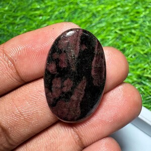 Pode incluir: Uma pedra preciosa polida de formato oval com uma base de cor preta profunda e manchas salpicadas de bord&ocirc; e vermelho escuro em toda a sua superf&iacute;cie lisa e reflexiva, exibindo uma textura de pedra natural e varia&ccedil;&otilde;es de cor distintas.