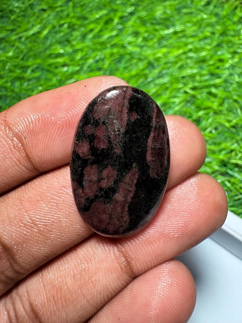 Pode incluir: Uma pedra preciosa polida de formato oval com uma base de cor preta profunda e manchas salpicadas de bord&ocirc; e vermelho escuro em toda a sua superf&iacute;cie lisa e reflexiva, exibindo uma textura de pedra natural e varia&ccedil;&otilde;es de cor distintas.