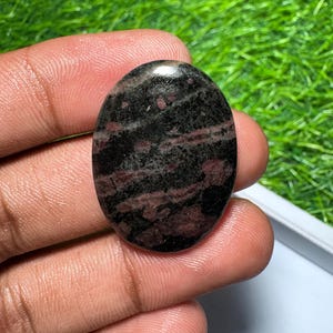 Pu&ograve; includere: Una pietra preziosa lucidata di forma ovale con una base di colore nero scuro e prominenti inclusioni minerali rosso intenso e marrone su tutta la superficie, caratterizzata da una finitura liscia, riflettente e lucida.